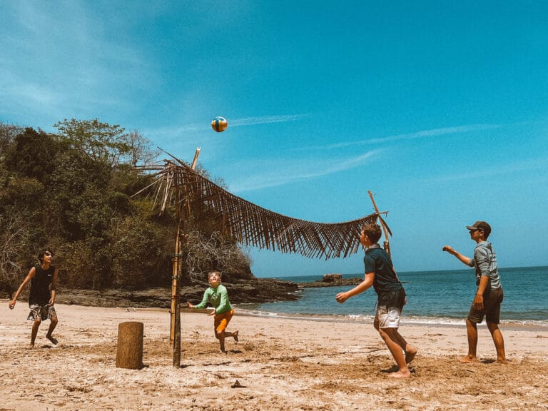Private Group Adventures 7 Kids playing beach volleyball on a tropical desert island with a makeshift palm leaf shade and ocean in the background.
