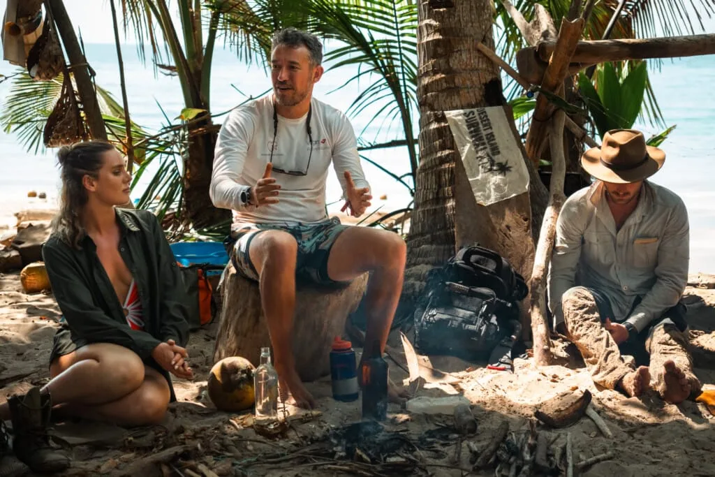 Survival experts discussing desert island survival techniques on a tropical beach.
