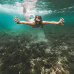 A person wearing goggles and a snorkel swims underwater above a seabed covered in algae and rocks, arms extended forward, pushing past any discomfort to explore the vibrant world below.