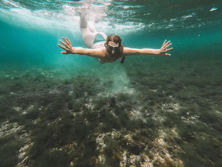 A person wearing goggles and a snorkel swims underwater above a seabed covered in algae and rocks, arms extended forward, pushing past any discomfort to explore the vibrant world below.
