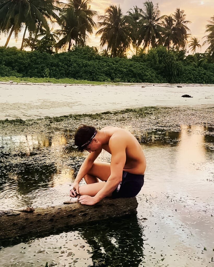 Desert Island Survival Adventure in the Maldives 10 A shirtless man sits on a rock by shallow water at the beach, focusing on something in his hands, as palm trees and a golden sunset set the scene for an Island Survival Adventure in the Maldives.