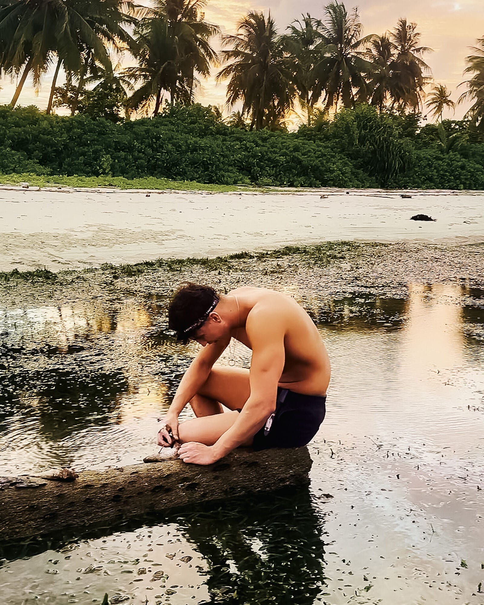 Desert Island Survival Adventure in the Maldives 25 A shirtless man sits on a rock by shallow water at the beach, focusing on something in his hands, as palm trees and a golden sunset set the scene for an Island Survival Adventure in the Maldives.