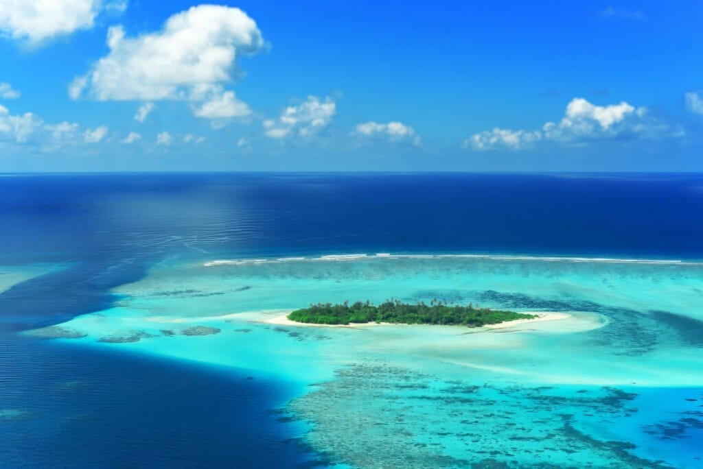 Desert Island Survival Adventure in the Maldives 14 A small, green island surrounded by turquoise water and coral reefs sits in the middle of a blue ocean under a partly cloudy sky, setting the perfect scene for an Island Survival Adventure in the Maldives.