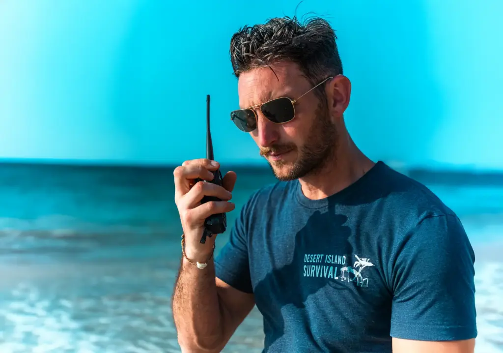Meet the Instructors: Firemakers, Fear-Tamers, Foundational Humans 4 A man wearing sunglasses, holding a walkie-talkie, at a beach, promoting desert island survival skills.