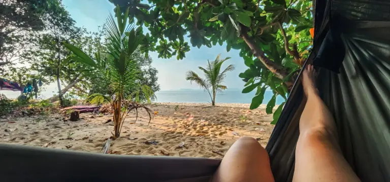 Lush tropical beach scene viewed from inside a hammock with person relaxing, surrounded by greenery, overlooking the ocean, perfect for desert island survival and outdoor adventure.