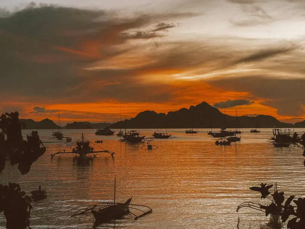 Boats anchored near a tropical island at sunset, showcasing a serene island survival scene with calm waters and vibrant sky colors.