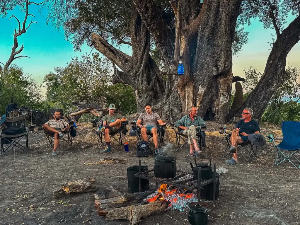 Botswana Kayak Bushcraft Safari in the Okavango Delta 9 A group of five men relaxing in foldable camping chairs around a campfire beneath a large, ancient tree on a deserted island, highlighting outdoor survival and survival skills in a wilderness setting.