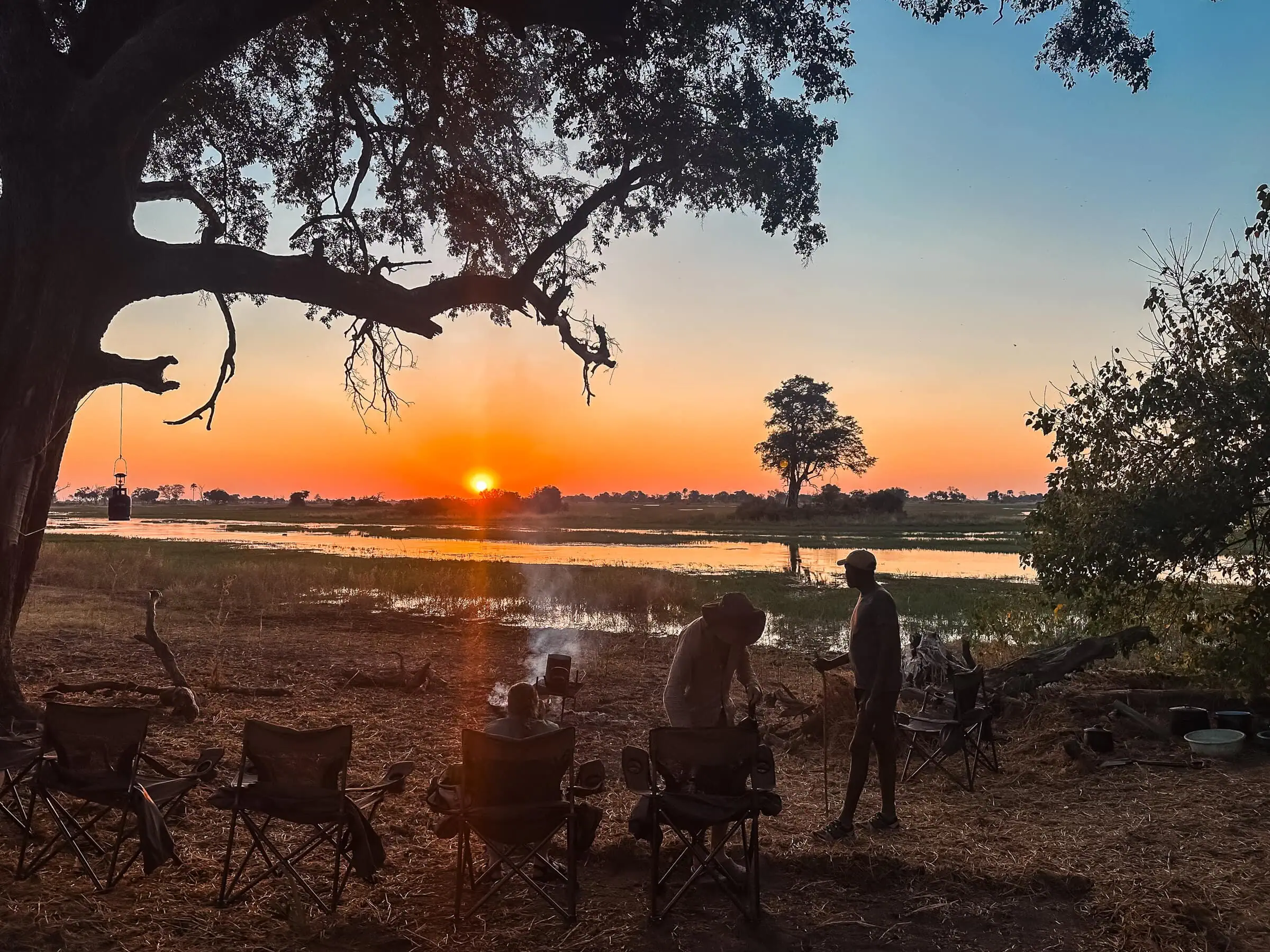 Botswana Kayak Bushcraft Safari in the Okavango Delta 23 Sunset over a tranquil river on a desert island, with people camping, cooking, and enjoying nature; perfect for desert island survival adventures and outdoor survival skills.