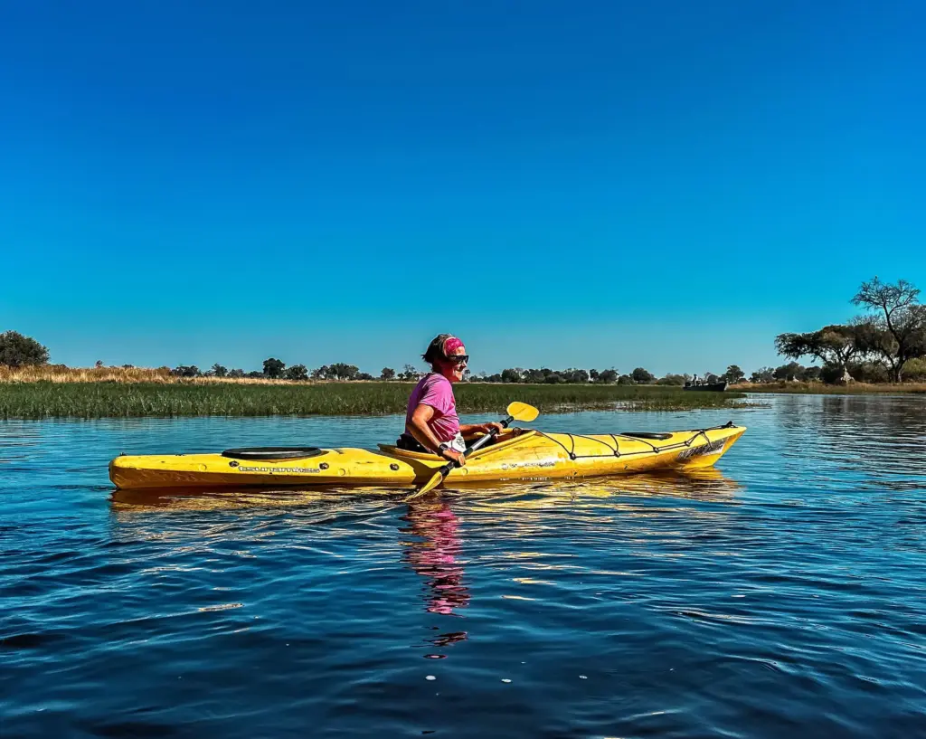 Botswana Kayak Bushcraft Safari in the Okavango Delta 10 Bright yellow kayak on calm water, featuring a woman in a pink shirt and headband paddling under a clear blue sky, perfect for island survival adventure and outdoor exploration.