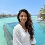A smiling woman with long wavy hair standing by an overwater bungalow pool in a tropical island resort.
