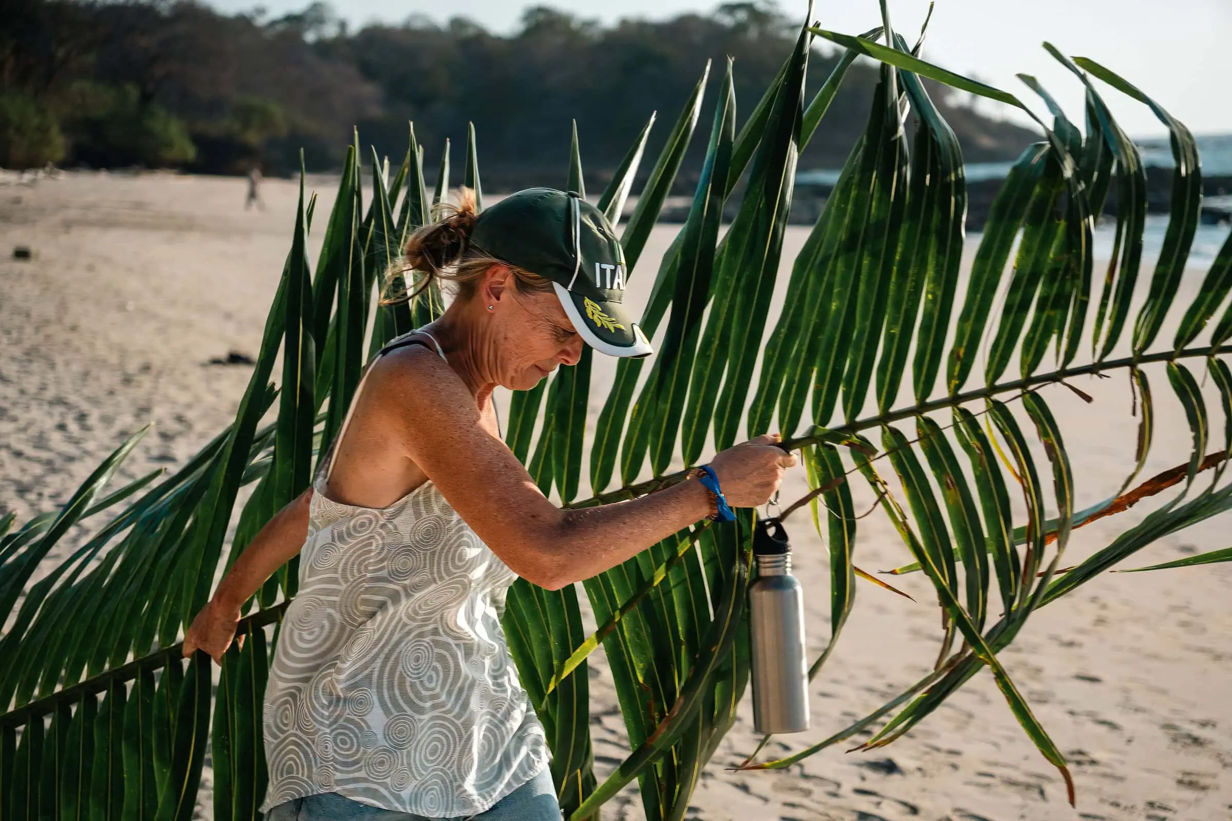 Lush green palm leaves on a sandy beach with a woman in casual clothing and a cap, holding a water bottle and exploring nature, highlighting survival skills on a desert island.
