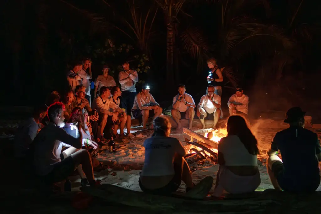 Survival skills on a desert island, group sitting around campfire at night, team building and adventure activities in an island setting, outdoor wilderness experience, survival training in tropical environment.