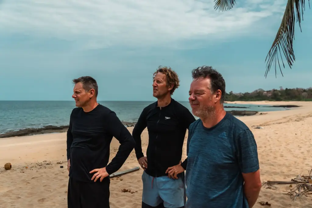 What Happens on a Desert Island (That You'll Carry Forever) 8 Men standing on tropical desert island beach, contemplating survival strategies, with ocean background and tropical palm leaves, emphasizing desert island survival skills and adventure in a remote location.
