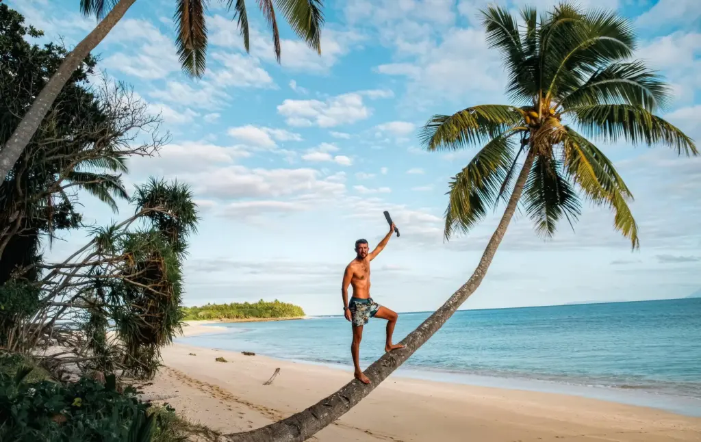Desert Island Survival Adventure in Tonga 5 A man climbing a leaning palm tree on a deserted tropical beach, with clear blue sky and calm ocean, showcasing survival skills on a tropical island for desert island adventure.
