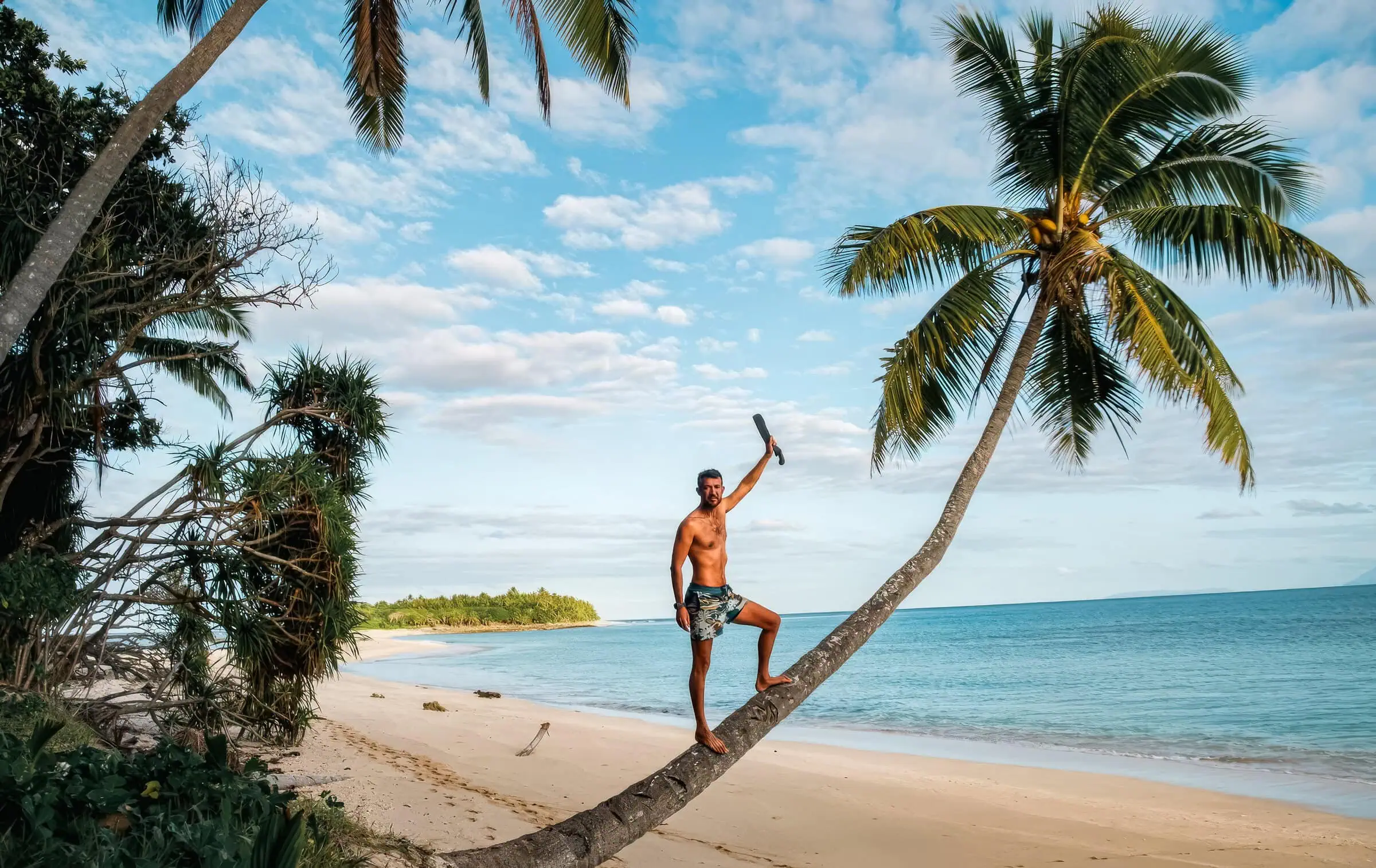 Desert Island Survival Adventure in Tonga 16 A man climbing a leaning palm tree on a deserted tropical beach, with clear blue sky and calm ocean, showcasing survival skills on a tropical island for desert island adventure.
