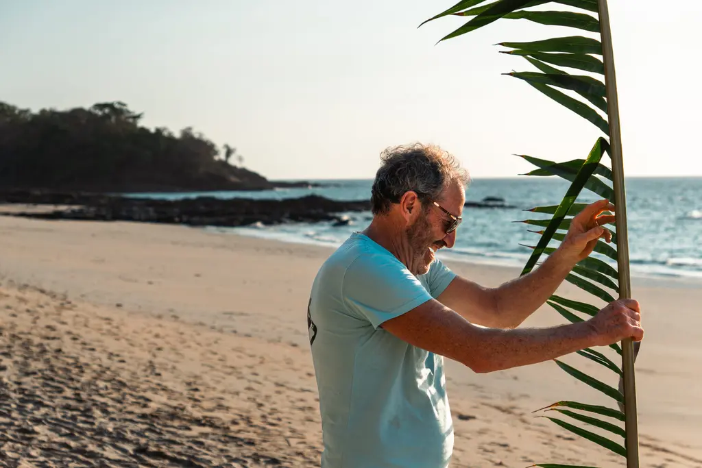 An older man wearing sunglasses and a light blue t-shirt stands on a sandy Panama beach, smiling as he holds a tall palm frond. The ocean and distant trees hint at island survival under clear skies.