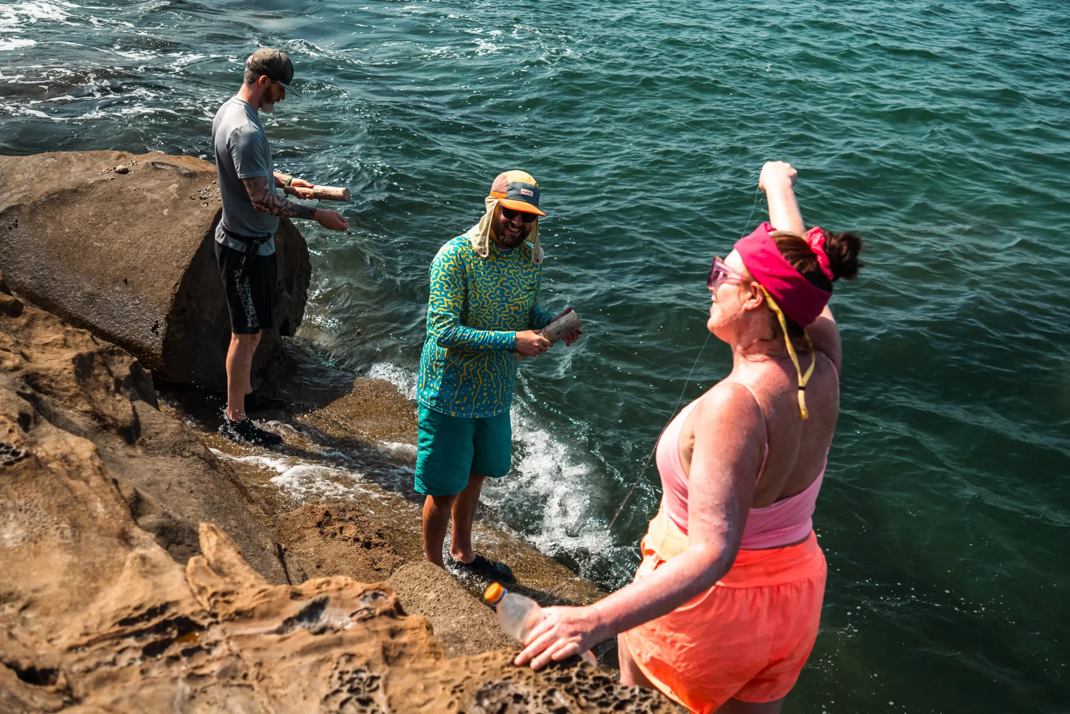 Three people on rocks near the ocean, one fishing and two talking.