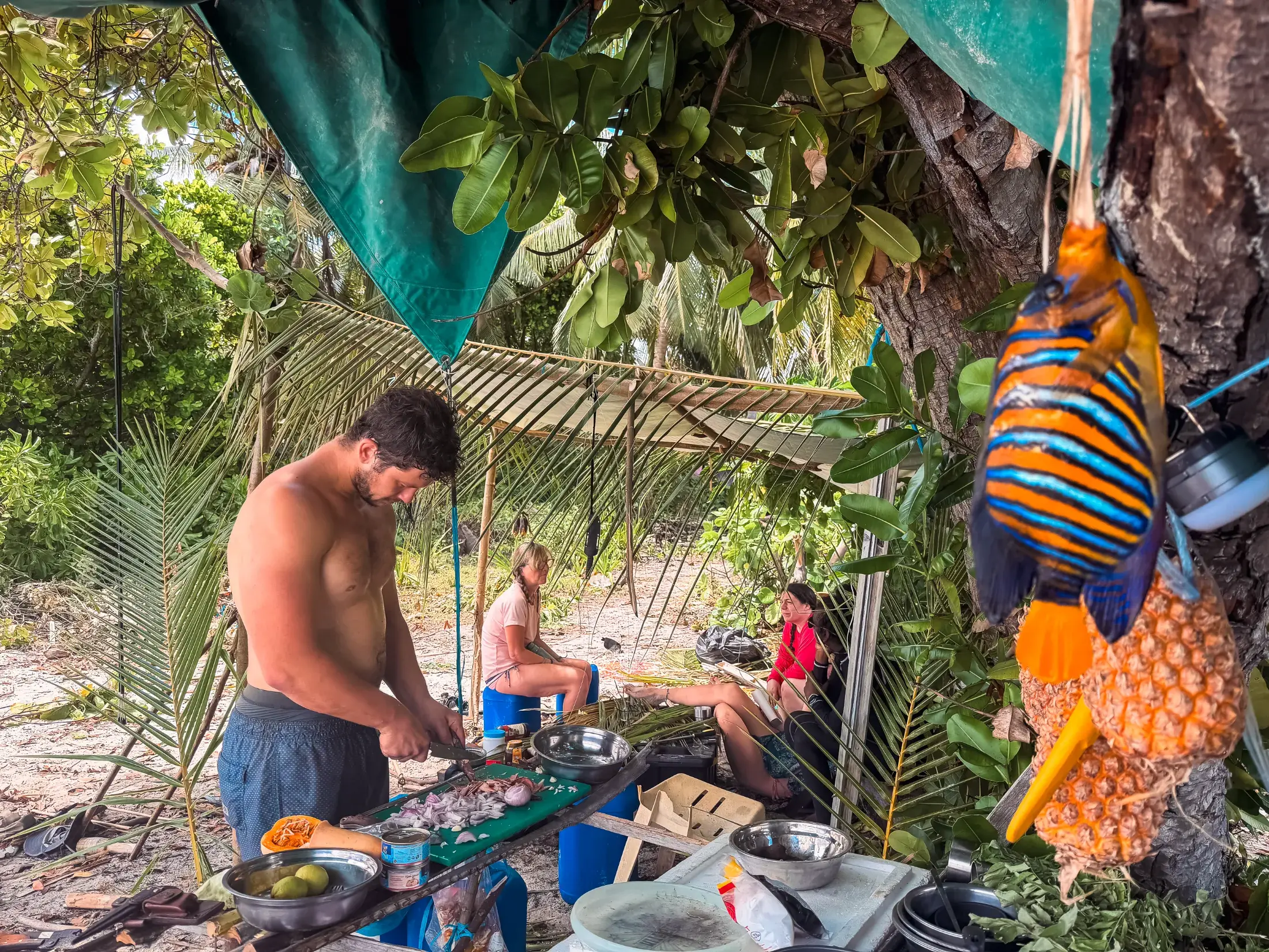 Man chopping vegetables during survival in the Maldives, with fish and pineapples hanging.
