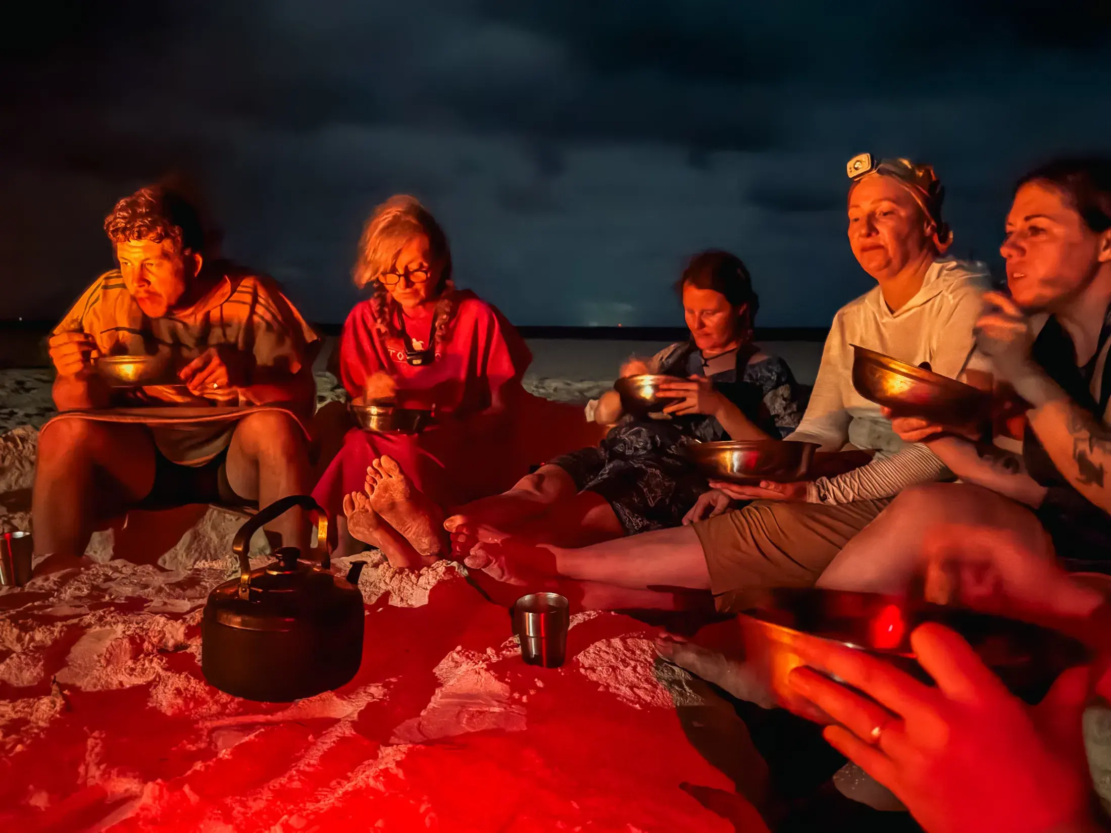 Group of people eating by a bonfire on a beach at night.