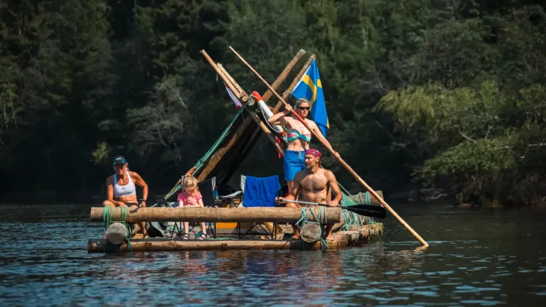 Family on a log raft with Swedish flag on a river in Sweden.