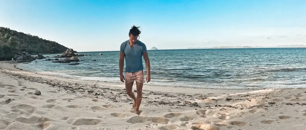 Man walking barefoot on a tropical beach with turquoise water and lush green hills in the background.