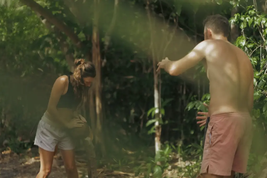Survival expedition: Woman crafting with man watching in jungle setting.