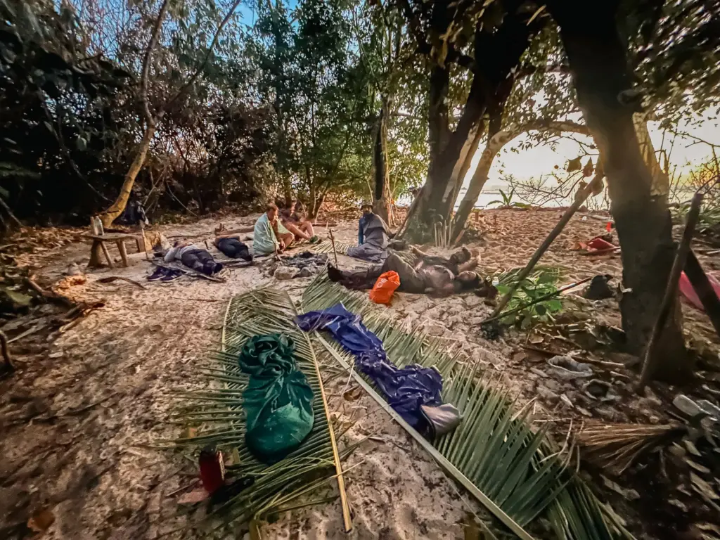 Survival expedition group relaxing on a sandy beach campsite under trees.