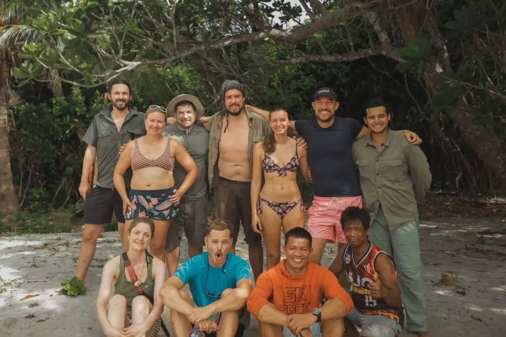 Group photo of survival expedition team on a sandy beach under trees.