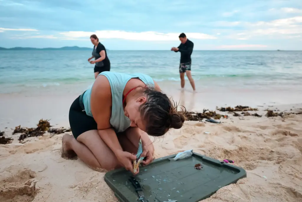 Woman preparing fish on beach during survival expedition with others in background.