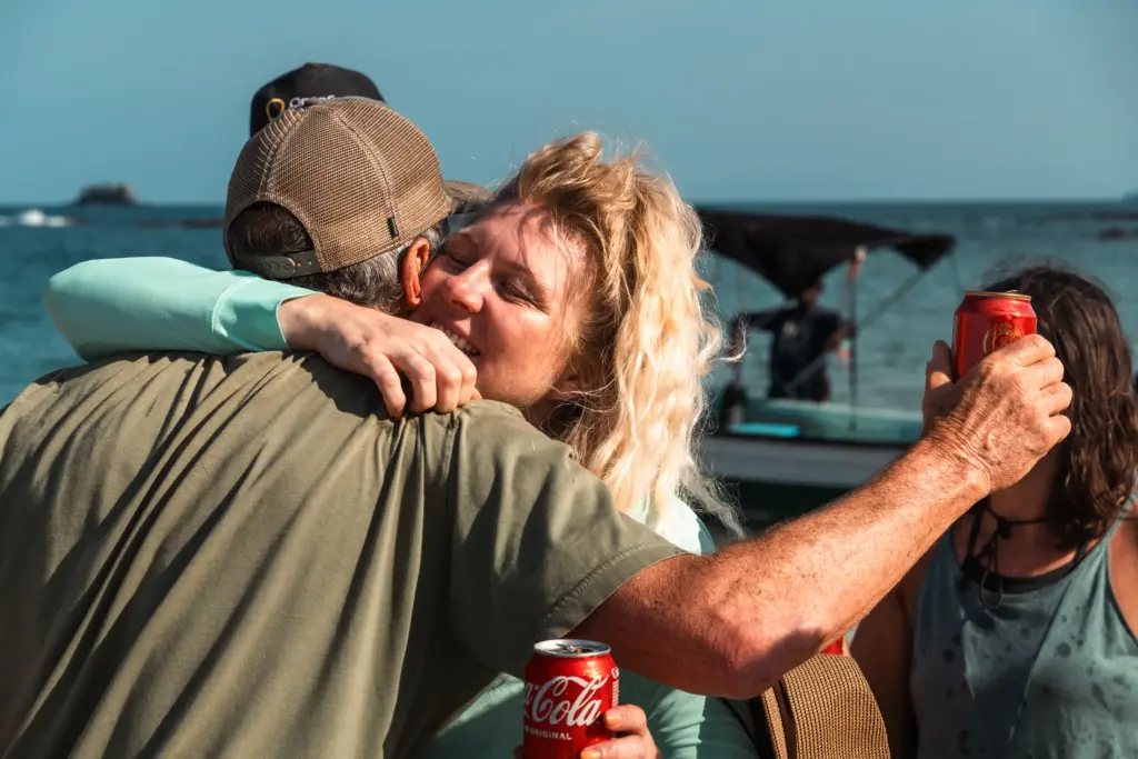 Woman hugging man on beach holding Coca-Cola can, boat in background.