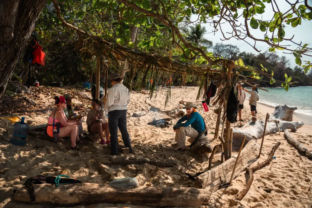 Desert island survival training: people gathered under makeshift shelter on sandy beach.