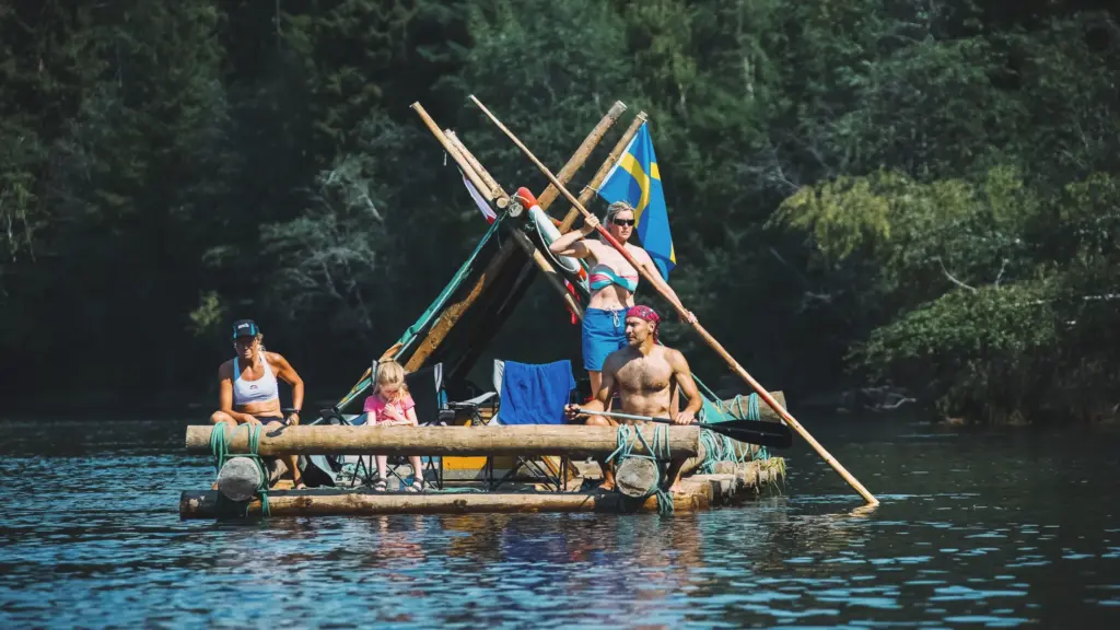 Family on a raft with Swedish flag, enjoying a sunny river adventure.
