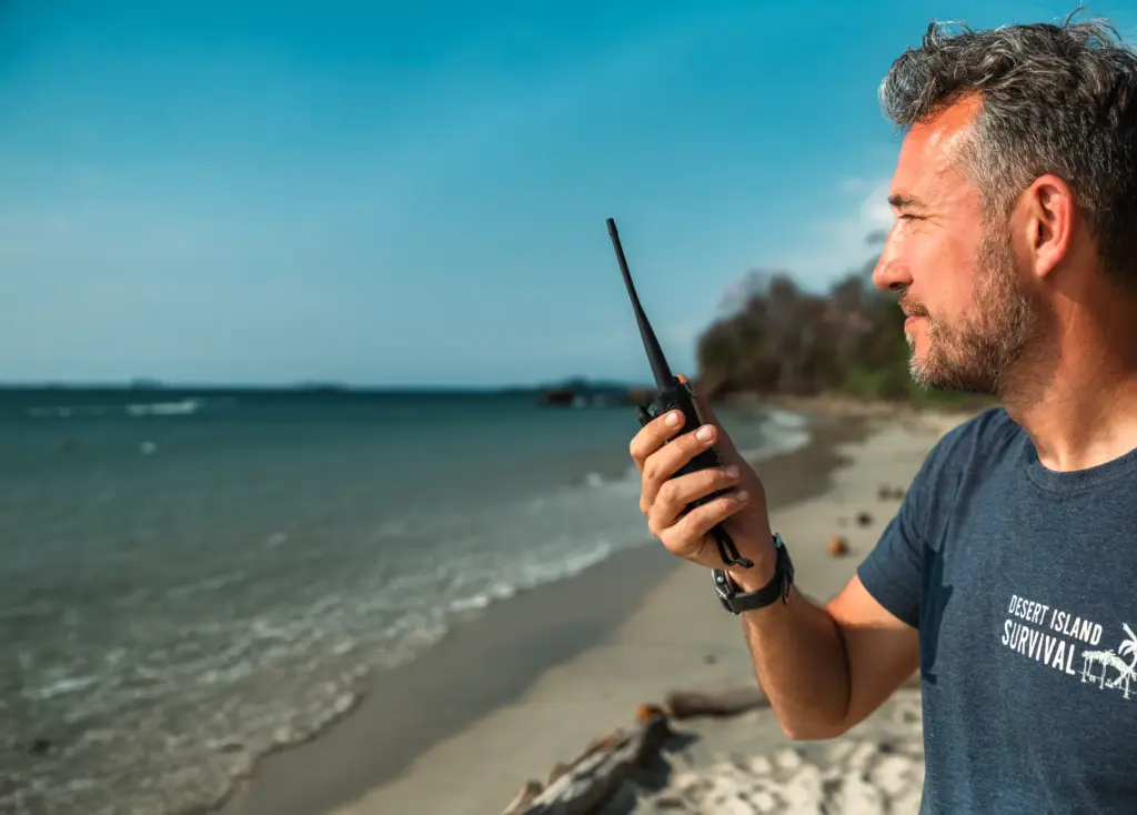 Man on beach holding walkie-talkie, wearing Desert Island Survival shirt.
