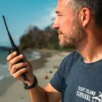 Man with walkie-talkie on beach. Desert Island Survival t-shirt visible.