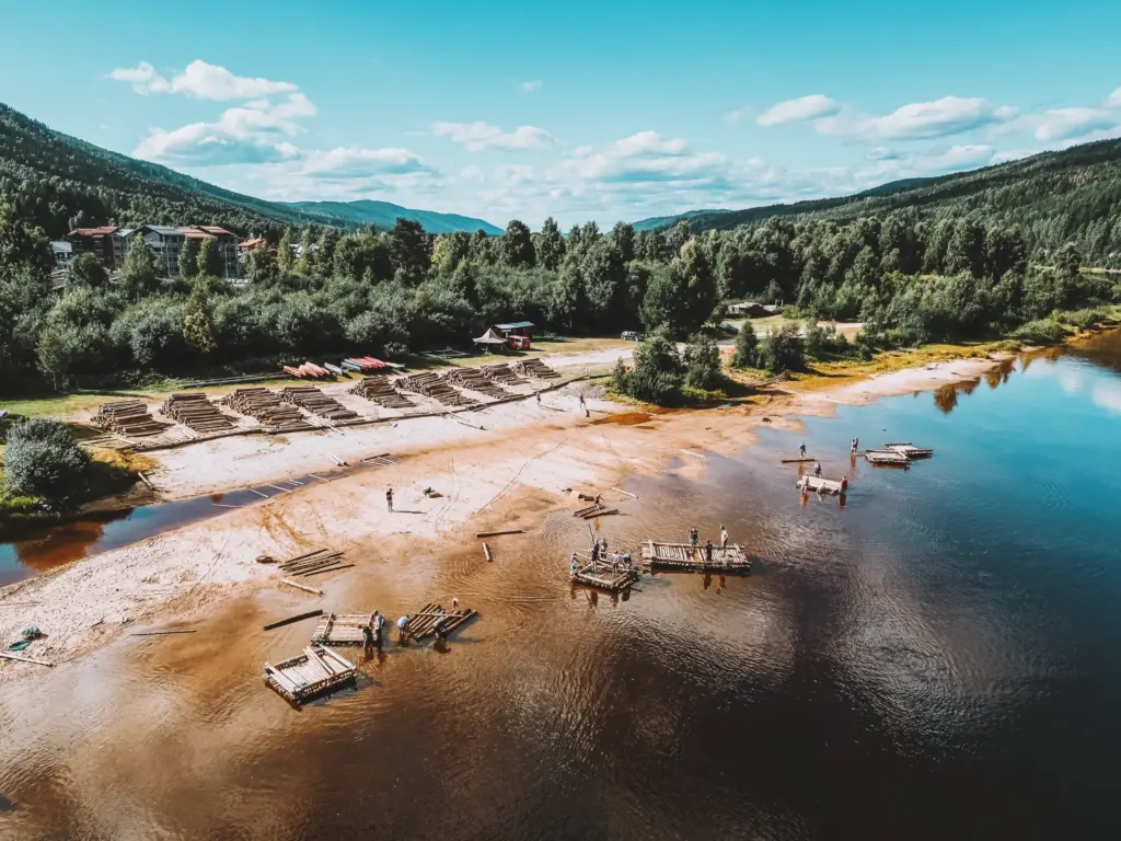 People building rafts on a sandy riverbank surrounded by forest and mountains.