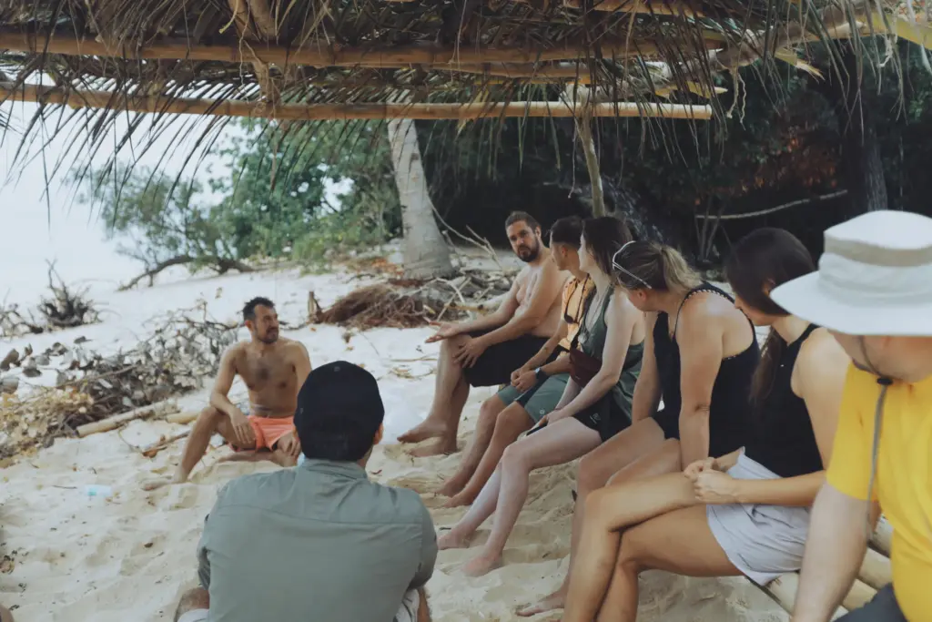 Group survival training session on a sandy beach under a makeshift shelter.