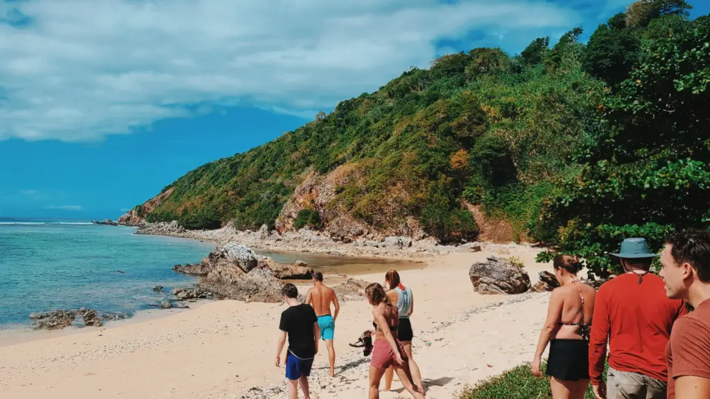 Group walking on tropical beach near jungle-covered cliffs, blue sky. Survival training context not directly visible.