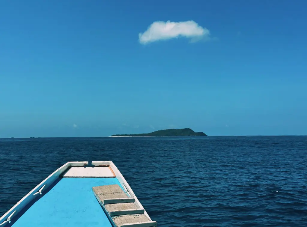The World Feels Dangerous Right Now. Here Is What That Actually Means for a DIS Expedition. Boat heading towards a tropical island under a clear blue sky with a cloud.