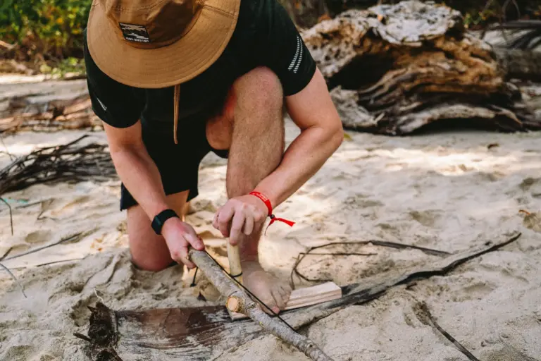 Man uses bow drill to start fire on sandy beach.