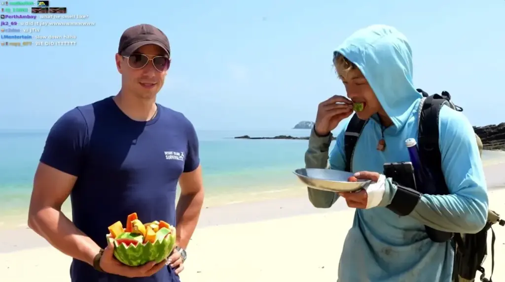 Two men on a beach, one eating from a watermelon bowl, wearing survival gear.