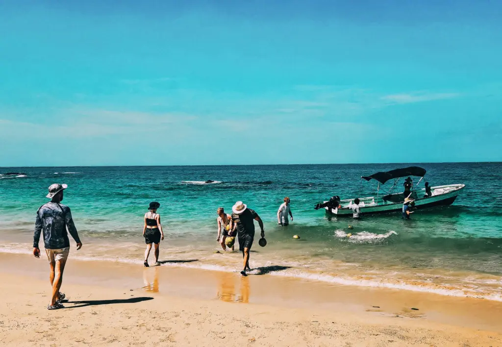 The World Feels Dangerous Right Now. Here Is What That Actually Means for a DIS Expedition. People wading near a boat on a tropical beach with turquoise water.