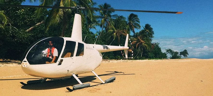 White helicopter on a beach with palm trees and a pilot inside.