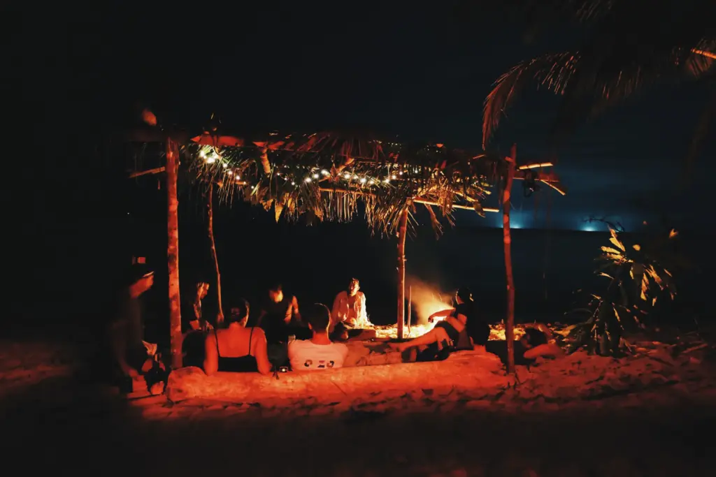 Beach bonfire with group under thatched shelter at night, survival expedition.