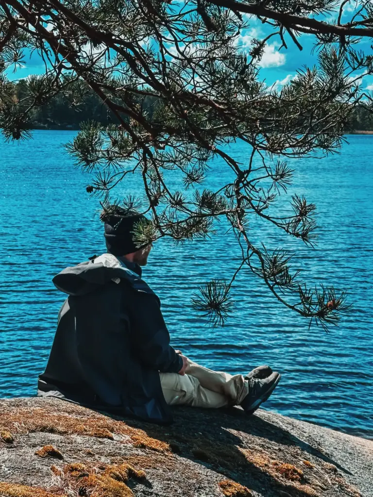 Man sits alone by lake in Sweden wilderness under pine tree branch.