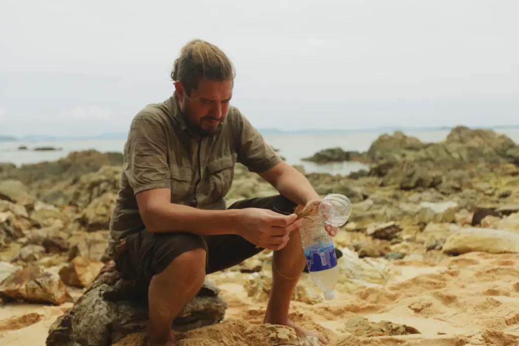 Man on beach starting fire with plastic bottle and kindling. Survival expedition.