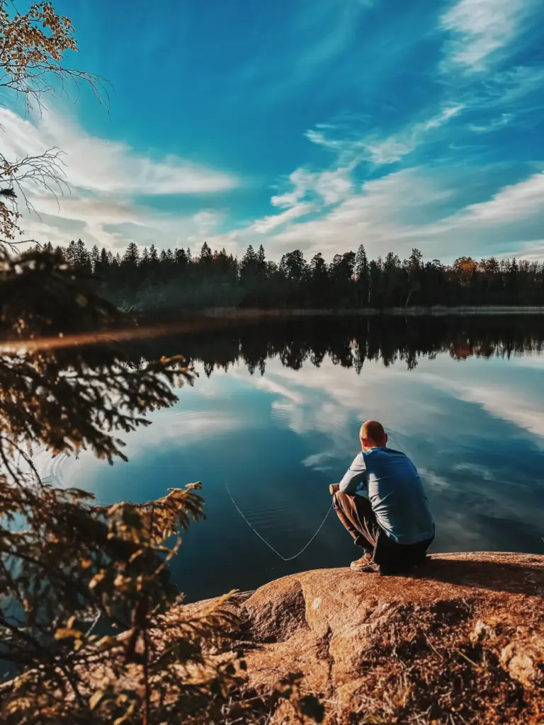 Man fishing alone on lake in Sweden wilderness. Calm water reflects trees and sky.