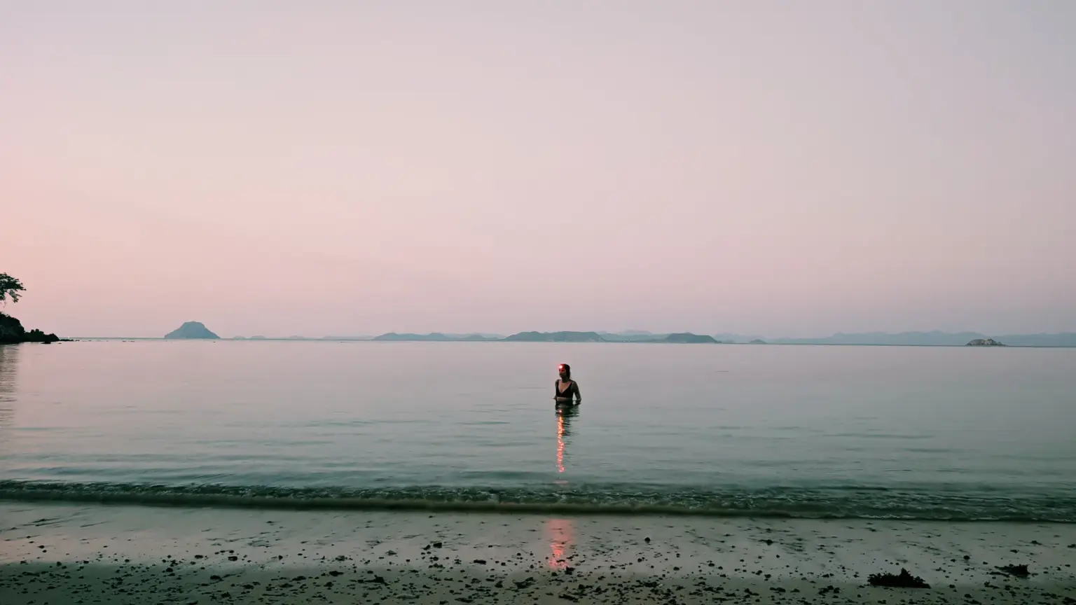 Person wading in calm ocean at dusk, with distant islands and a tranquil, survival expedition setting.