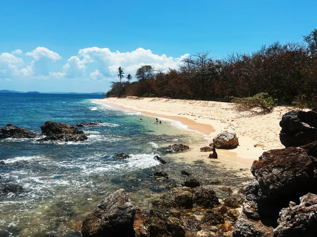 Tropical beach scene with turquoise water, sandy shore, rocks, and people swimming under a blue sky with clouds.
