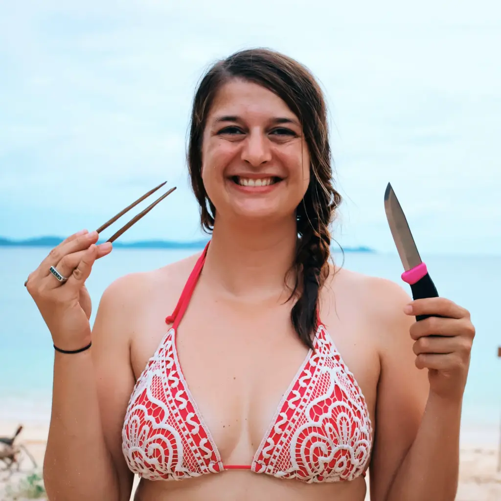 Woman on beach with chopsticks and knife, smiling. Survival expedition skills.