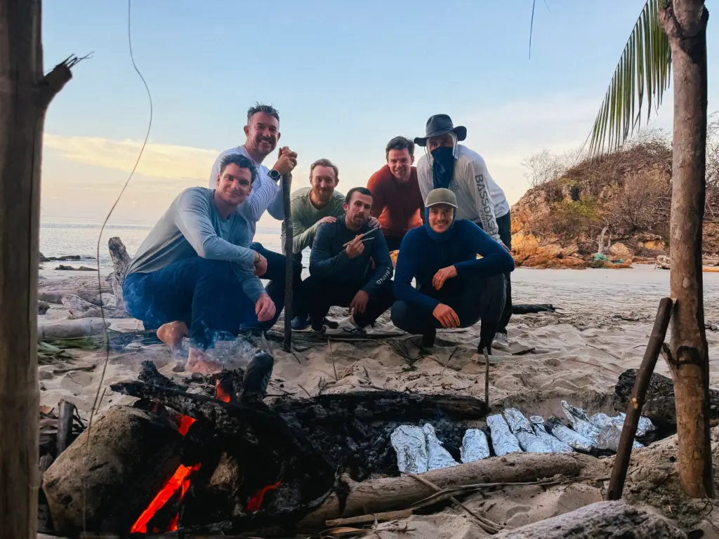 Group of men around a campfire on a beach, cooking food in foil.